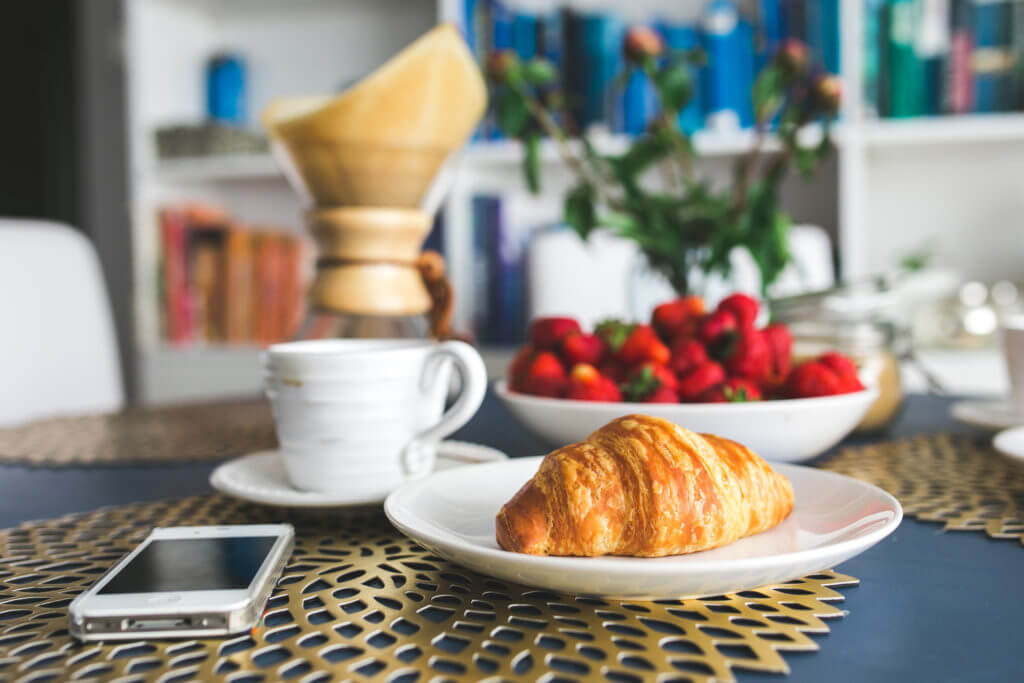 A phone sitting on a kitchen table next to a croissant, coffee cup, and a bowl of strawberries