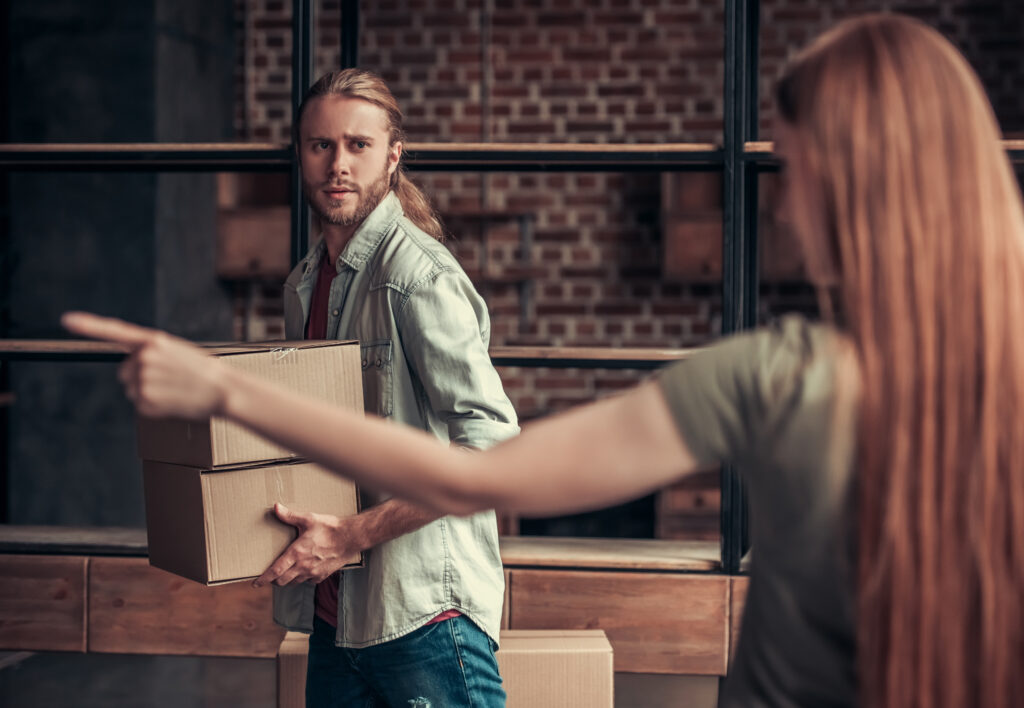 A woman pointing to the door while a man carries two cardboard boxes 