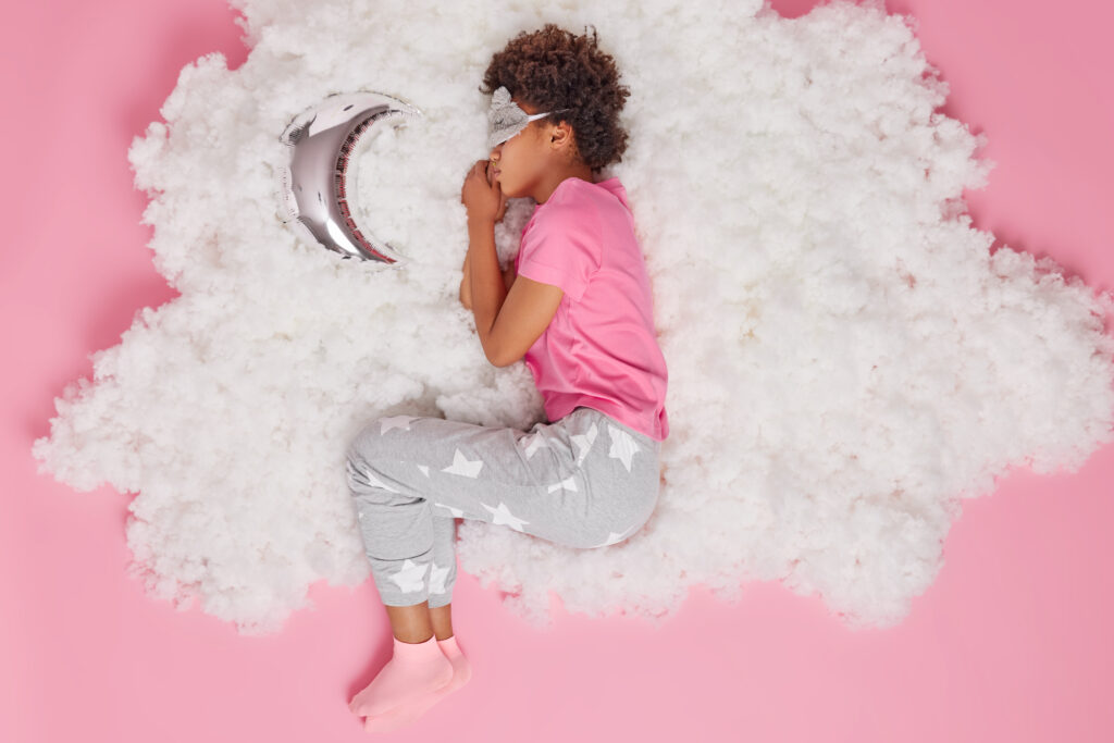 Overhead shot of relaxed calm child wears pajama and sleeping mask on eyes sleeps well during night sweet dreams poses on white cloud 