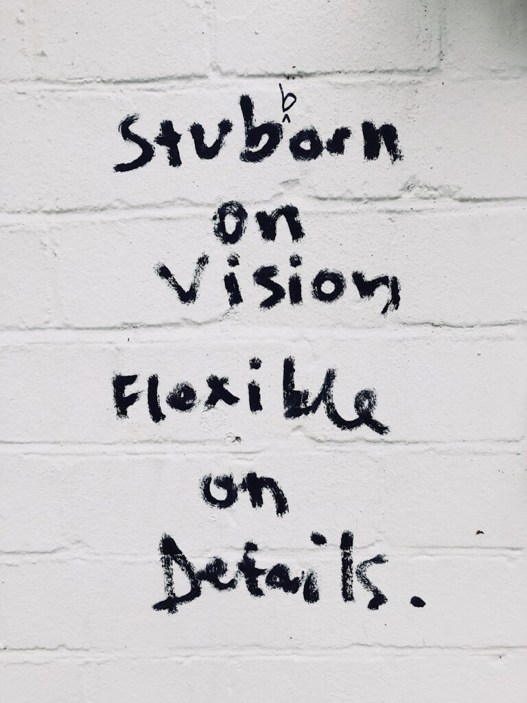 A white brick wall with the words "Stubborn on vision flexible on details" written on it
