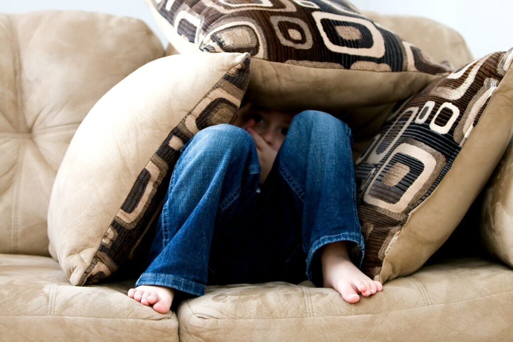 A child sitting on a couch, hiding under a pile of pillows. 