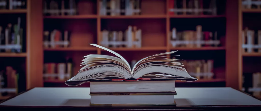 Three books sitting on a table in a library.  One of the books is open.  