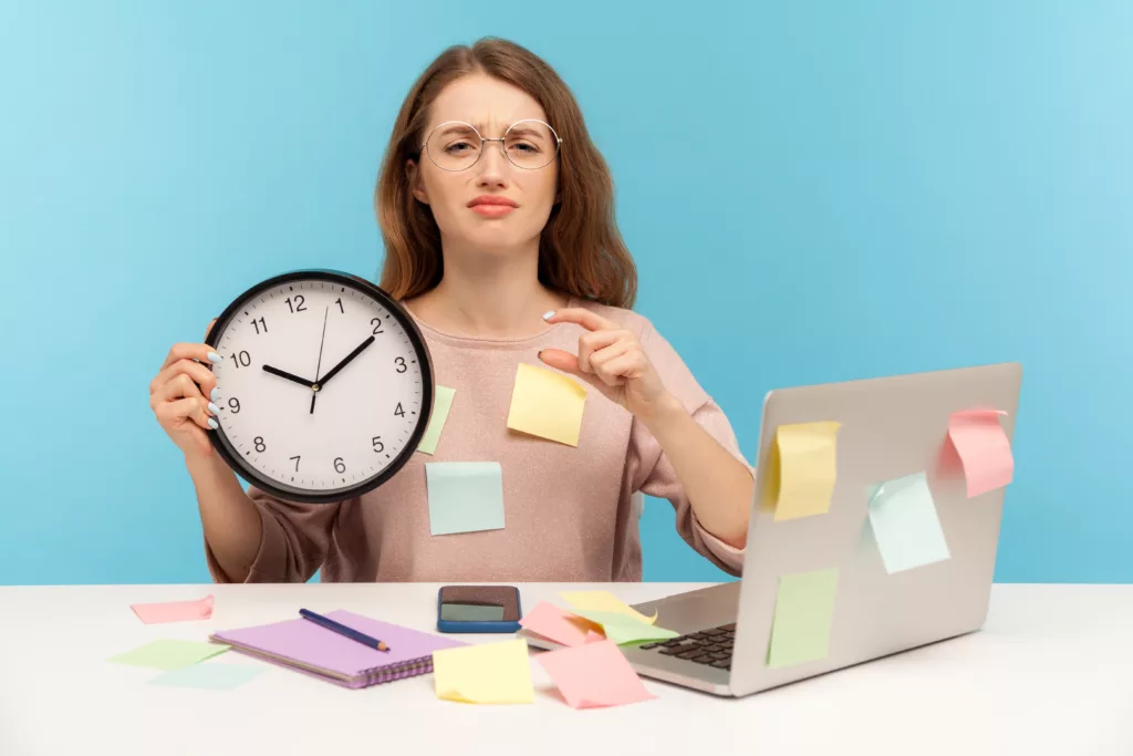 A woman sitting behind a laptop holding a clock.  She is covered in post it notes.
