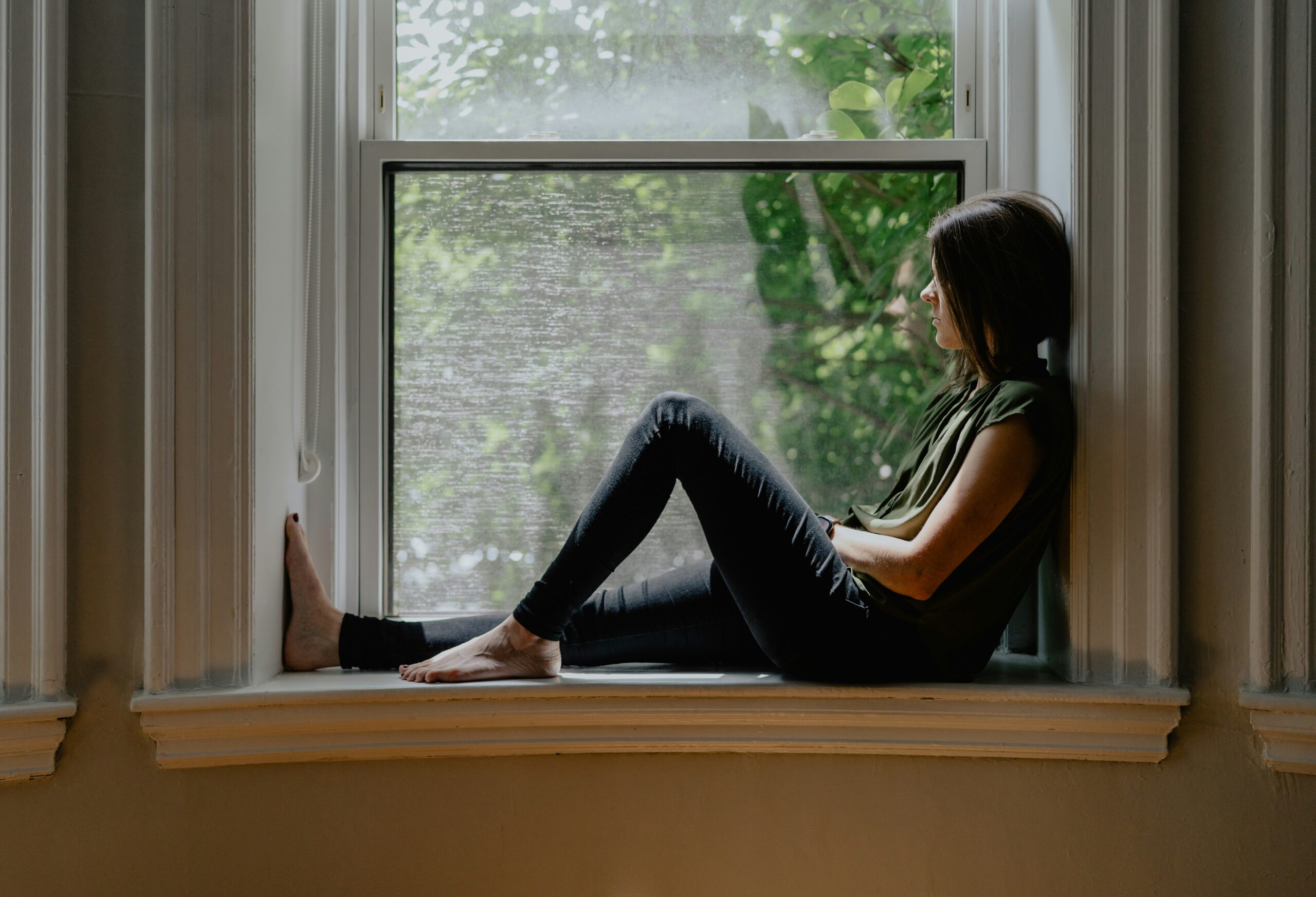 A woman sitting quietly by a window thinking about life coach or therapist
